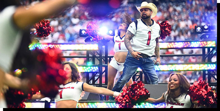A man, Louie The Singer, dressed as a cowboy, with a straw hat on stage next to dancers/ cheerleaders holding a microphone.